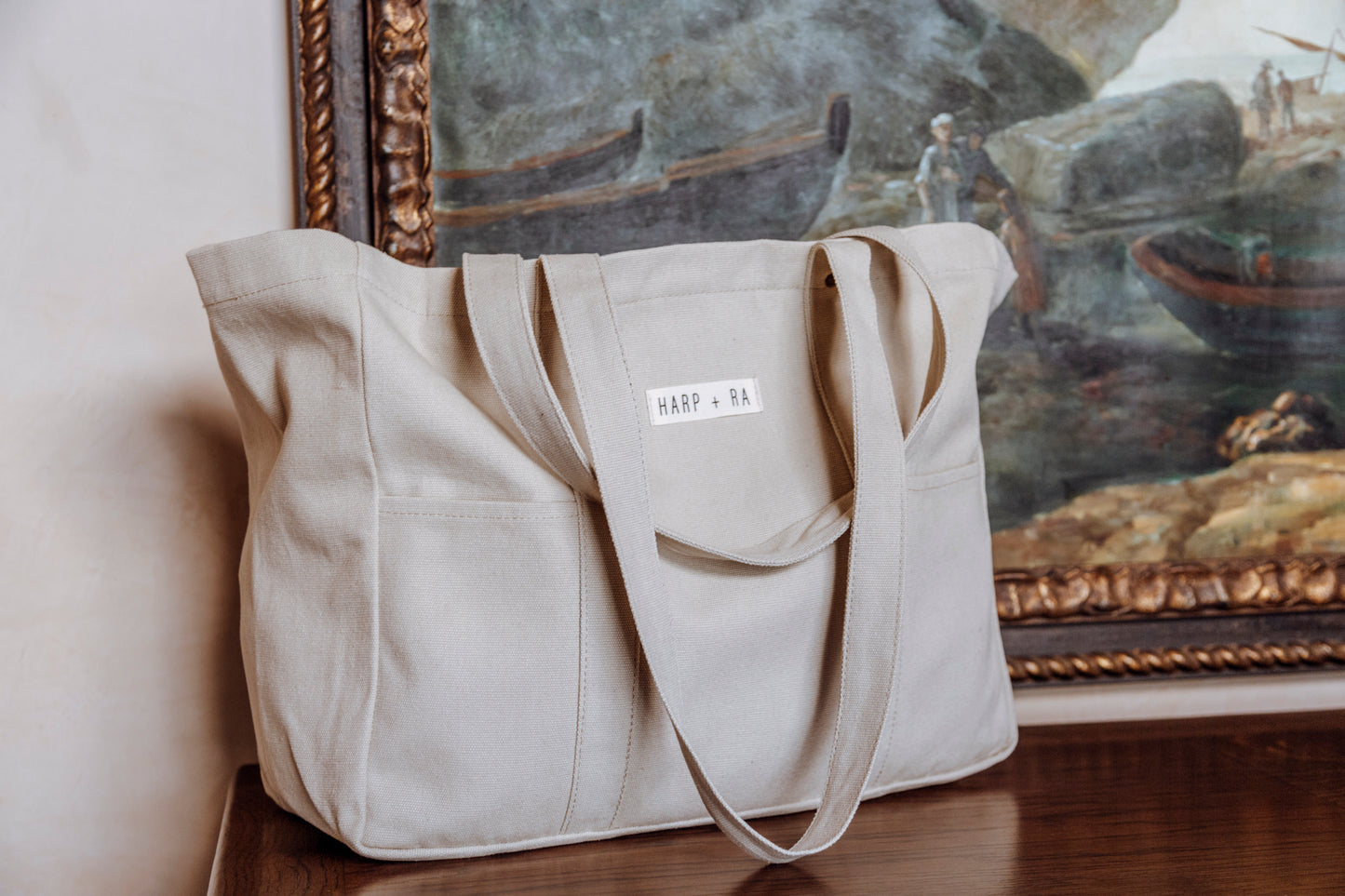A large beige womens tote bag sits on a cabinet in front of a painted picture, long shoulder handles hanging to the front of the bag with phone pockets